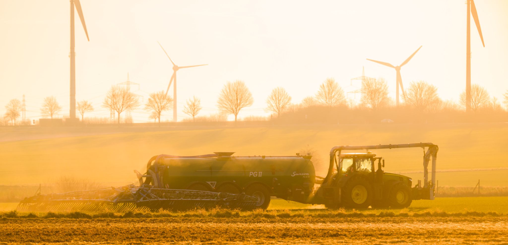 Mit einem Traktor wird bei Sonnenaufgang ein Feld mit Gülle gedüngt.