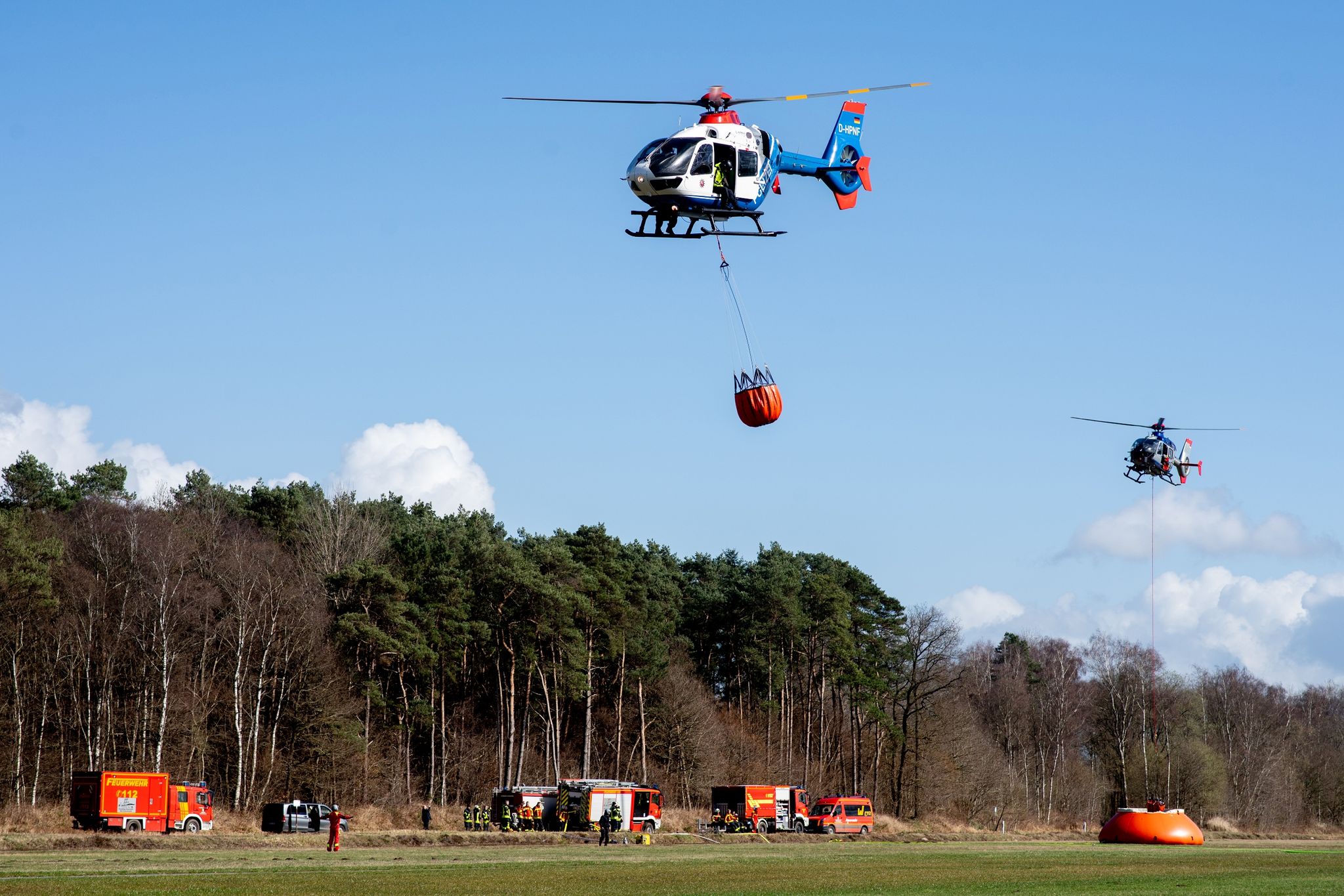 Hubschrauber der Polizei aus Niedersachsen (vorne) und aus Sachsen fliegen mit Löschwasserbehältern, sogenannten Bambi Buckets, über einen Flugplatz.